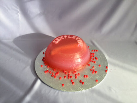 A pink jello cake on a silver platter with red berries scattered on the platter. The cake has a ceramic dildo inside it, and has the word SEXPLETIVE decorated on the top. The platter stands on a white sheet. 