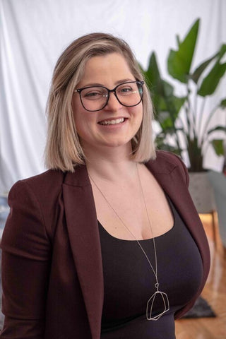 A femme presenting person with short blonde hair, black glasses and a brown blazer with a black top stands in front of a office background with a plant visible.