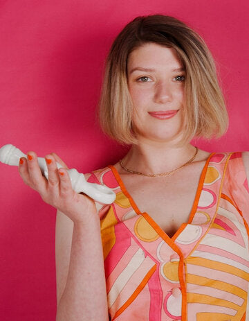 A femme presenting person with short blonde and brown hair stands in front of a hot pink background. They hold a white ceramic sex toy and are wearing a pink and orange top which is vibrantly coloured and patterned.