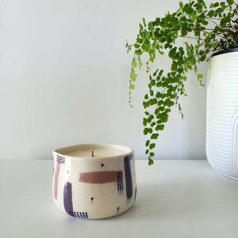 A ceramic massage candle in a purple pattern stands on a white table against a white wall. A bright green fern flows from a pot on the right.
