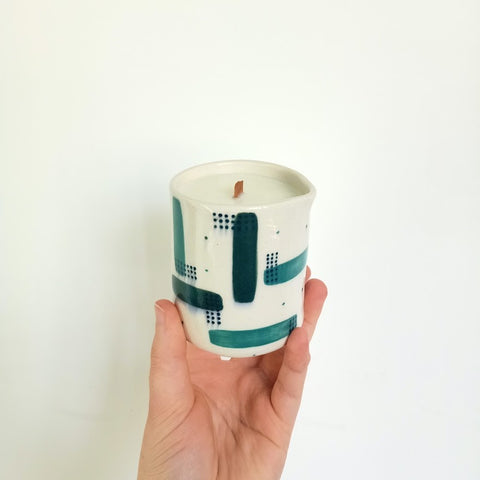 A massage candle in a green and teal pattern is held by a hand in front of a white wall. The candle is tilted forward so you can see the wooden wick in the centre of the candle.
