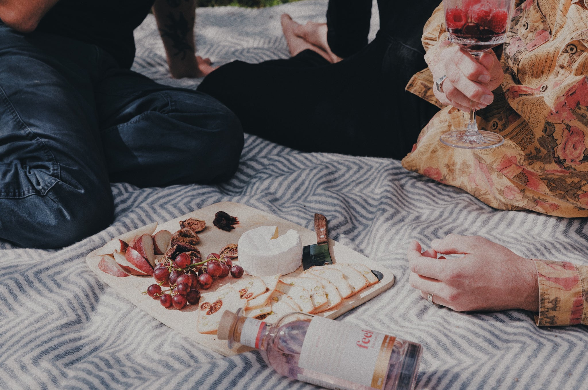 A bottle of Rouse being enjoyed on a picnic rug by two people while eating a cheese board