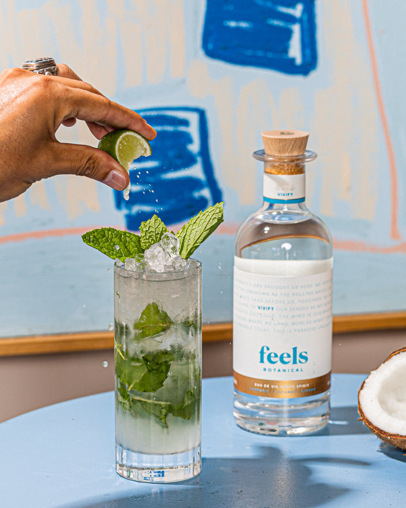 A hand squeezing a wedge of lime into a tall cocktail glass with mint and lime next to a bottle of Vivify.