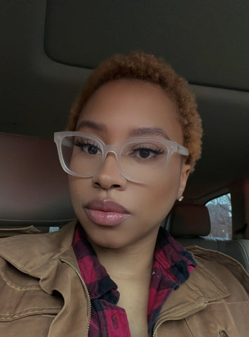 light skin woman in car with glasses. Tan jacket red & black checker shirt