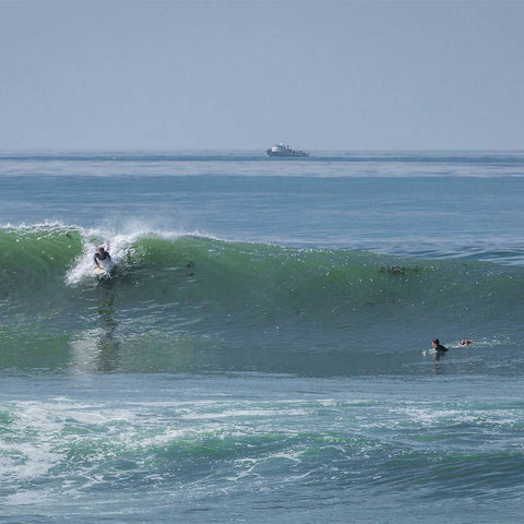 Two men are out surfing.  One man is surfing a large wave, while the other is watching him from the side.  There is a commercial fishing boat in the distance.