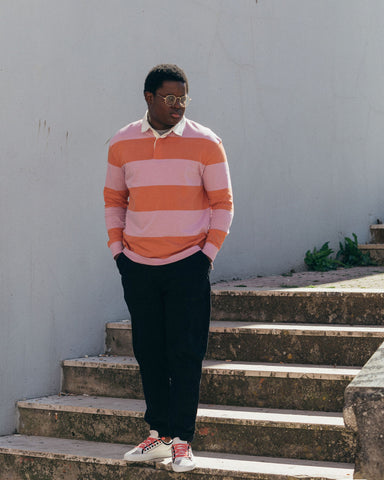 Ein Mann in einem pink-orangefarbenen Pullover und einer schwarzen Hose, der auf einer Treppe steht und Diverge sneakers trägt, hebt die soziale Wirkung und die maßgeschneiderten Schuhe durch das imagine-Projekt hervor.