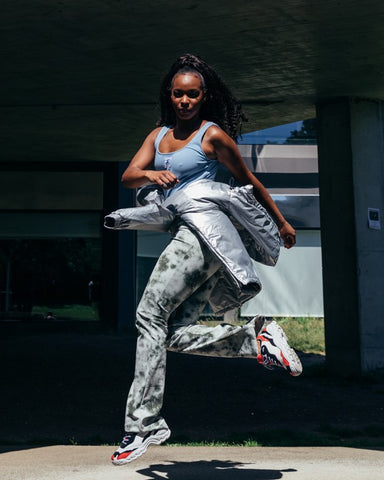 A woman in a blue top and grey pants jumping, wearing diverge sneakers, promoting social impact and custom shoes through the imagine project.