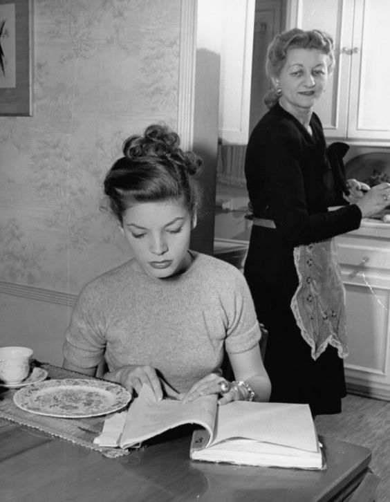 Lauren Bacall with her mother Natalie. Bacall reads a book at a table whilst her mother looks over her shoulder.