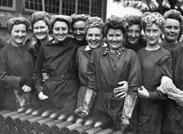 A group of female factory workers in WW2. All are wearing overalls, protective clothing, and have their hair up using headscarves and turbans.