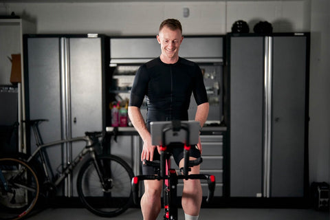 a man in dark cycling gear uses an indoor cycling trainer in a grey garage