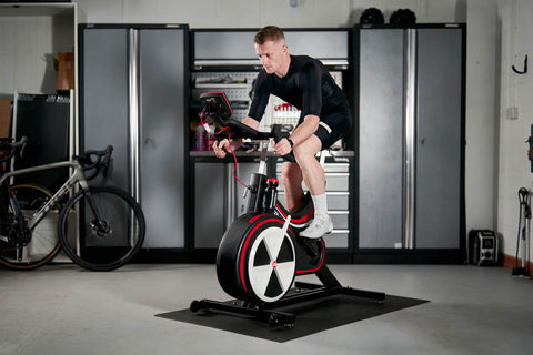 a male cyclist in a black cycling gear trains in a garage on an air bike