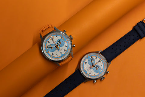 One watch with the Papaya Orange strap on the Pilote Racing Chronograph watch face wrapped around an orange cylinder. Another Pilote Racing Chronograph watch face with the Shelby Blue strap lead flat beside it on a solid orange backgroud.