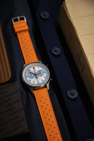 Papaya orange strap on the Pilote Racing Chronograph watch face. The watch is placed on a dark blue fabric backgroud with nutural tones accessories placed on the perimeter.