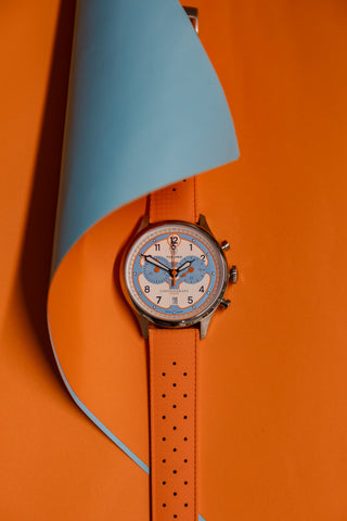 Papaya orange watch strap on the Pilote Racing Chronograph watch face. The watch is placed on orange paper with a blue curl of paper over the top.