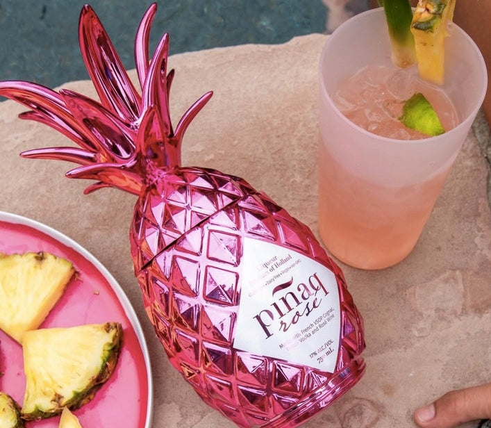 Pink bottle of Pináq Liqueur Rosé lying down with a tropical class of Pináq Rosé to the right and a plate of pineapple to the left.