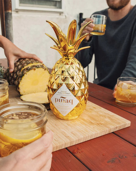 pinaq liqueur pineapple shaped screw top bottle on a wooden board next to an open pineapple and a person holding a cocktail