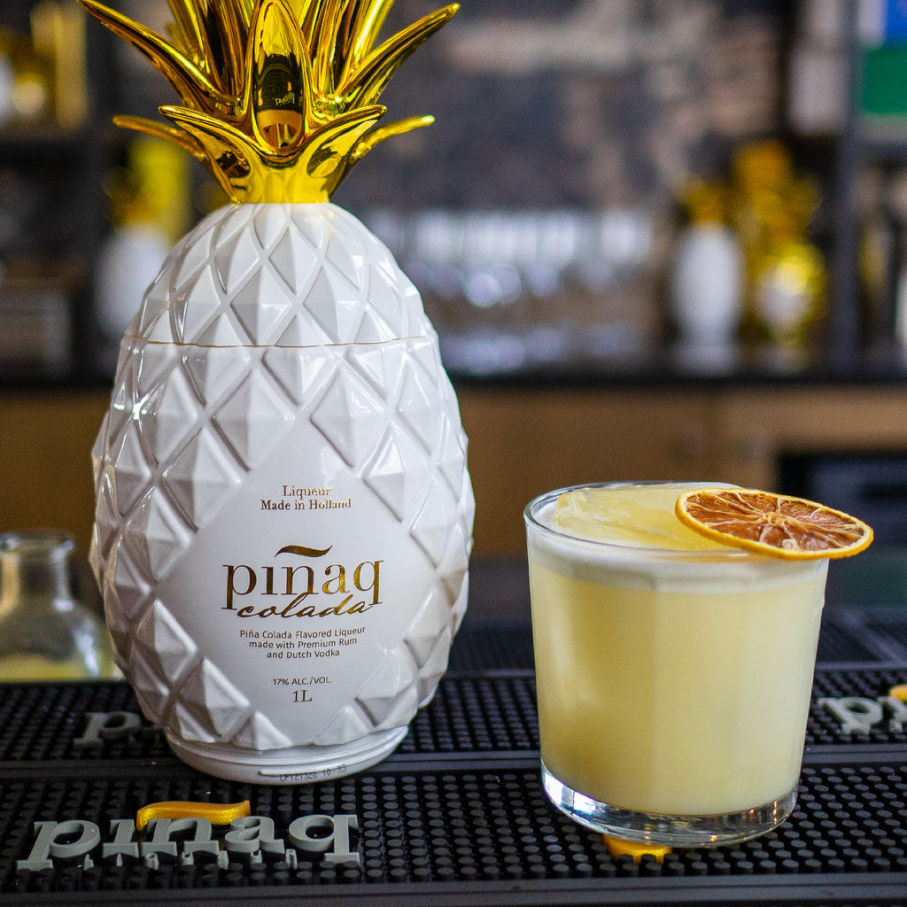 Pineapple shaped white and gold bottle of Pináq Colada with glass of Pinaq Colada cocktail to the right on a table.