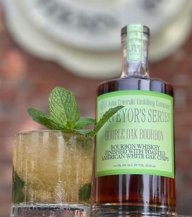john emerald purveyors series double oak bourbon next to a cocktail with herb garnish