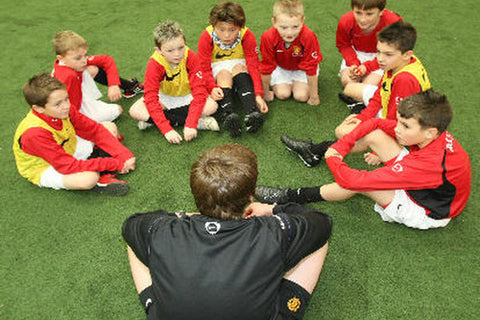 football coach talking to young players who are sat in ground in front of him. 