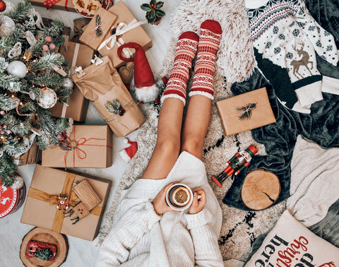 A woman with holiday socks and a mug sits amid wrapped gifts and a Christmas tree
