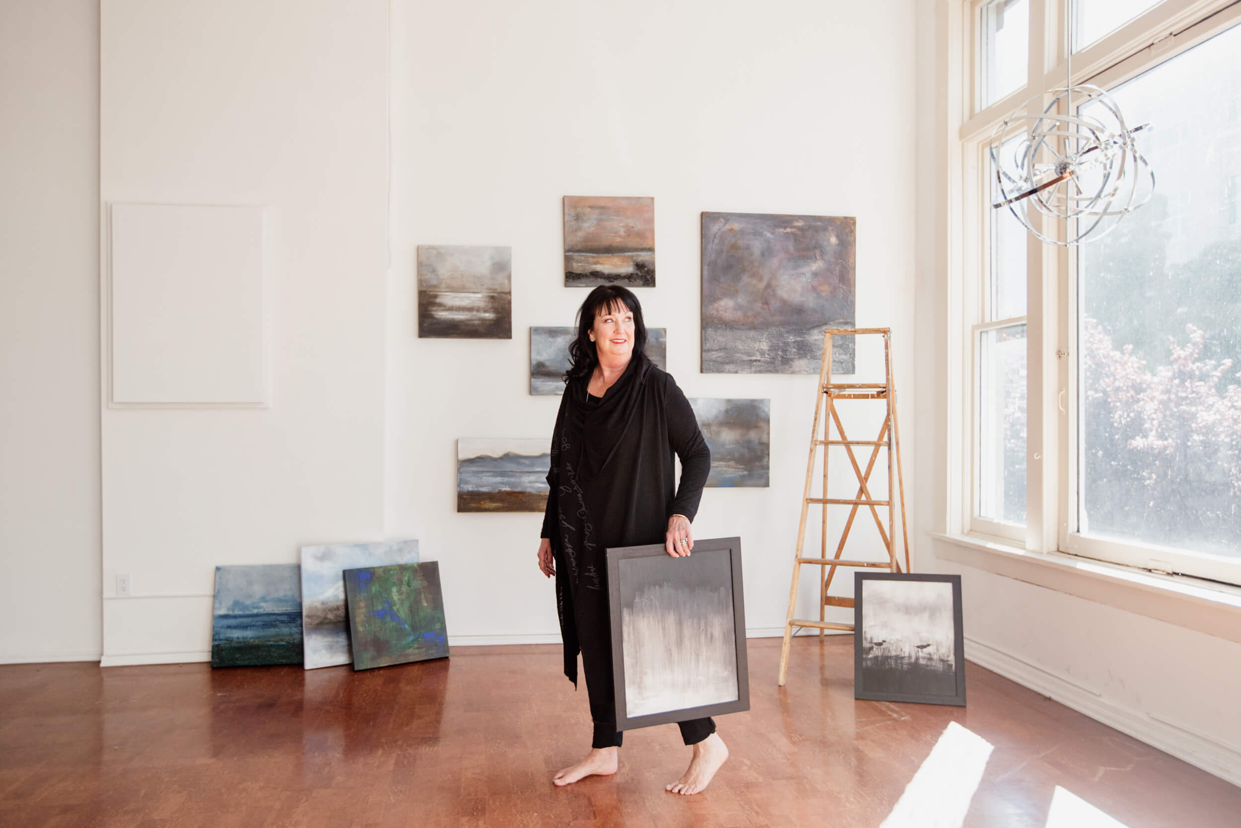 Artist Donna Anderson in her studio holding a painting standing in front of her wall of paintings.