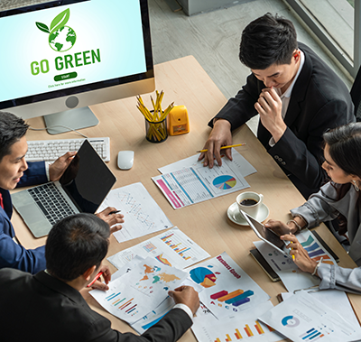 4 people sitting at a table with computers and papers, a large monitor in the back reading "go green."