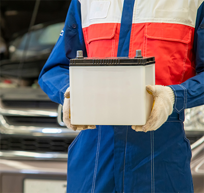 Person in a blue, orange, and white jumpsuit holding a white car battery standing in front of a car with a popped hood.