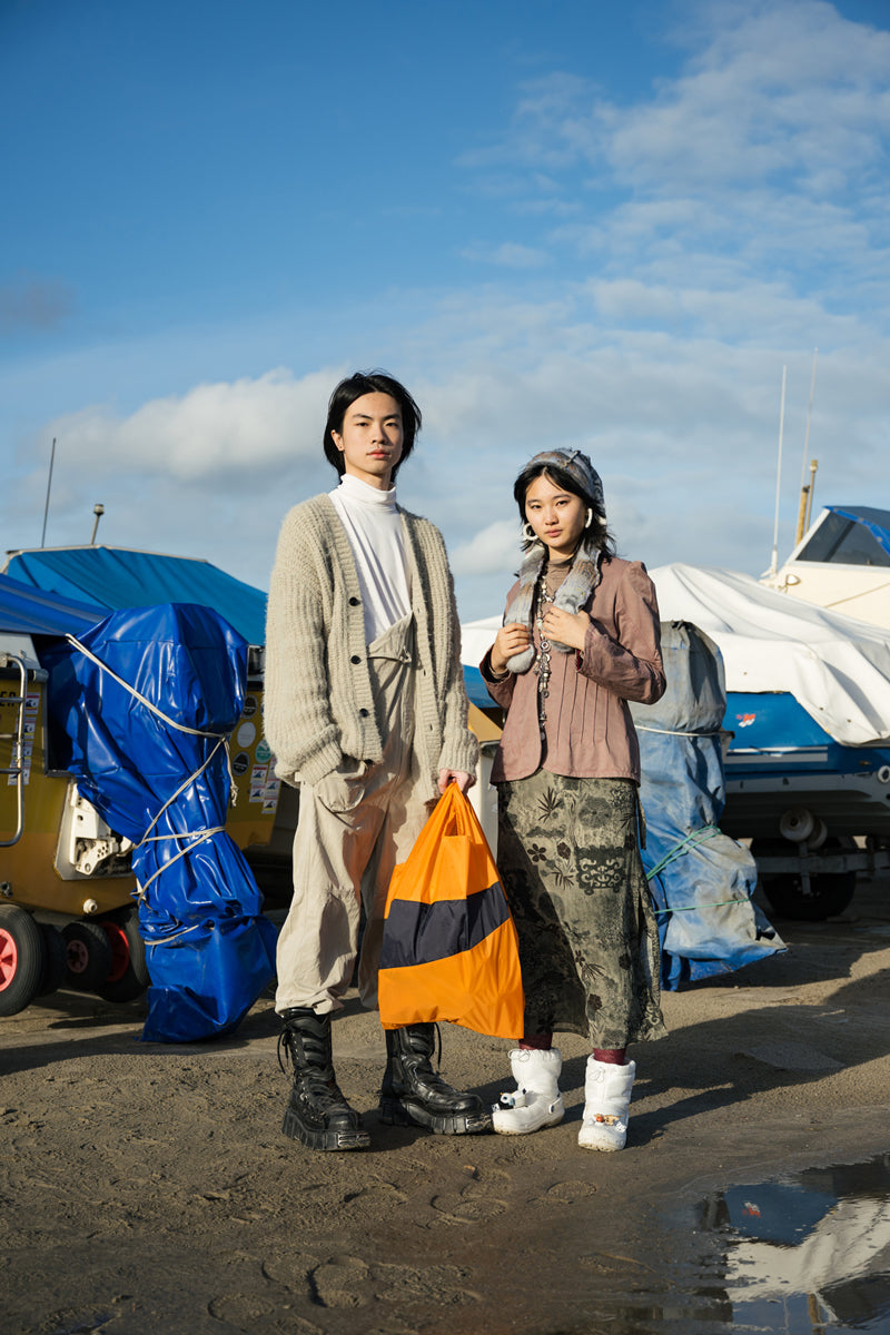 two models standing on the beach in front of fishing boats, with a bright orange shopping bag.