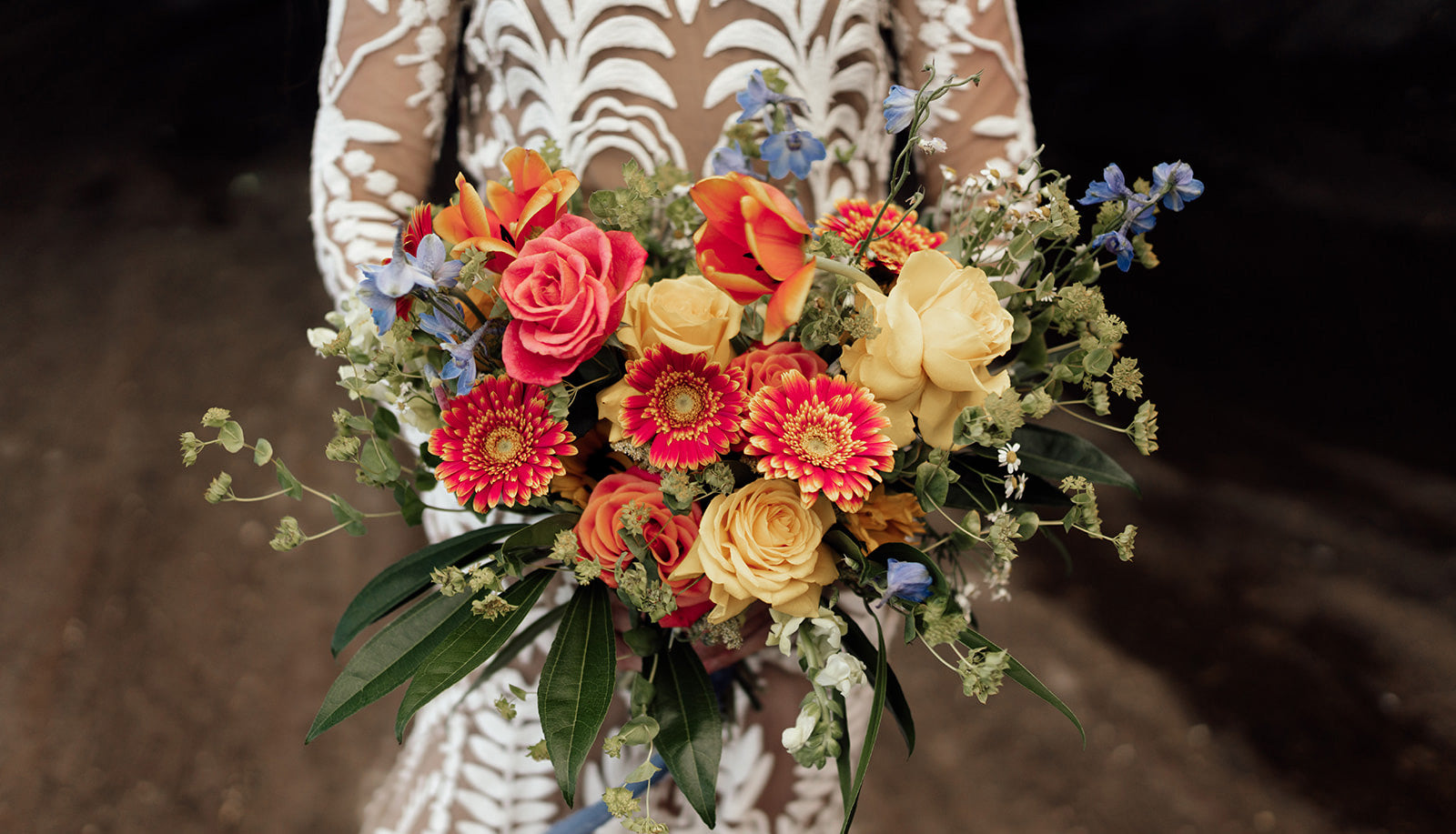 Bride holding a brightly colored bouquet of gerbera, roses and delphinium tied with blue ribbon.