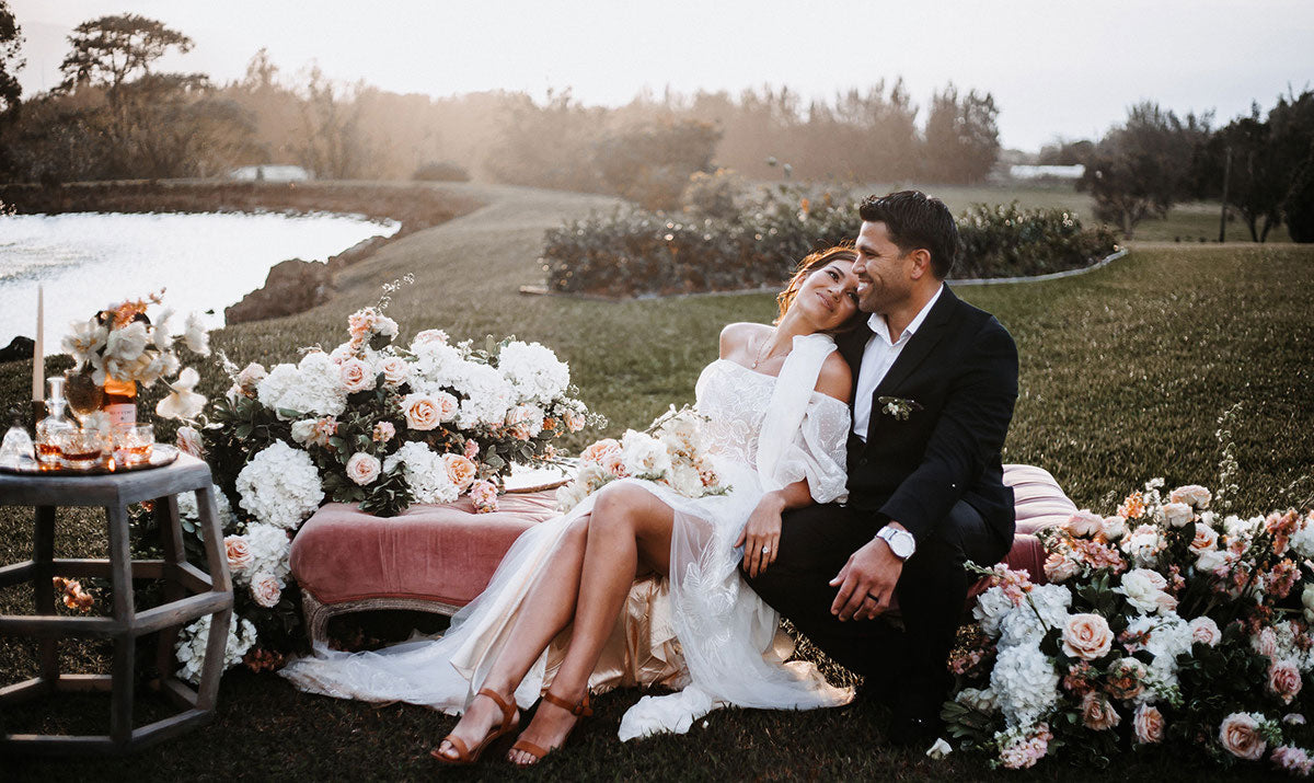 Bride and groom lounging on an ottoman surrounded by flowers outside