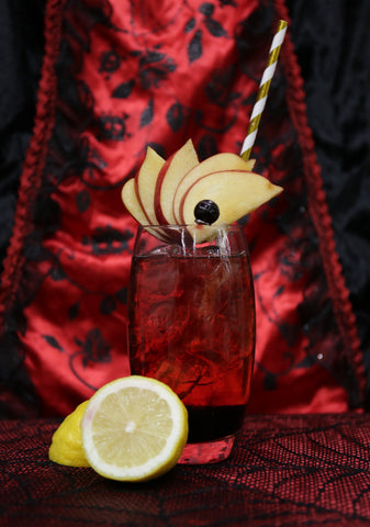 Photo of a tall cocktail glass with the Chimayó cocktail in. Set against a red with black lace backdrop, garnished with apples slices and a cut lemon sits in front of the glass. 