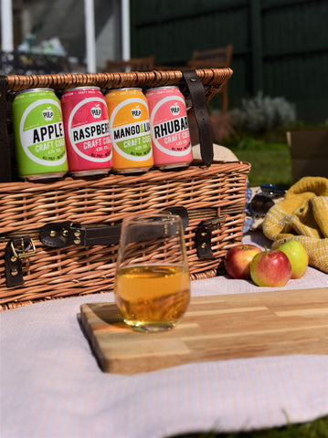 Photo of 4 cans and one glass of Pulp cider, and  picnic basket on a sunny day.