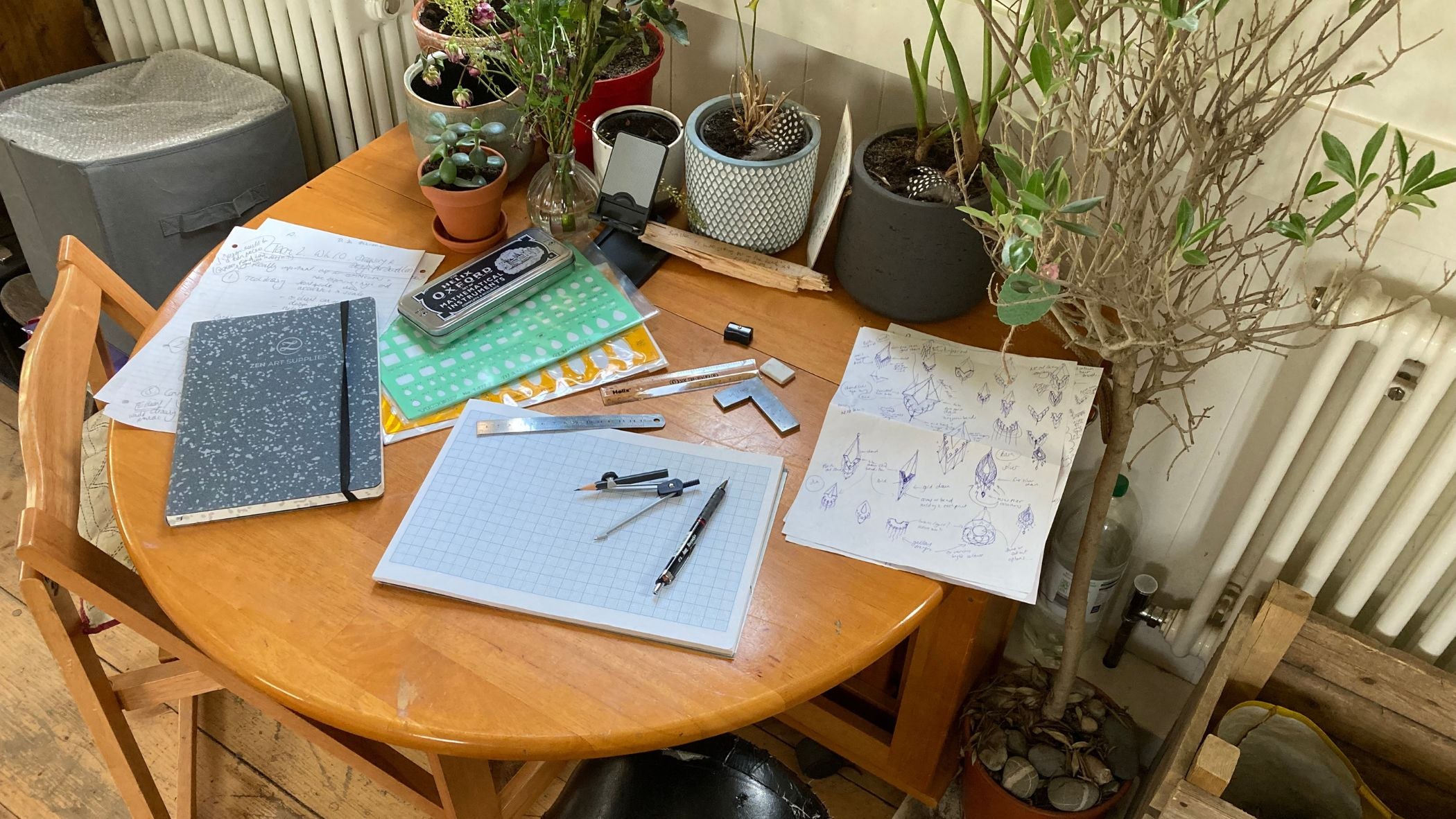 Kitchen table with notepad, pens, compass, other drawing equipment and sketches to one side.