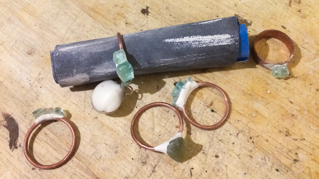 Copper rings attached to bits of broken glass and seaglass with epoxy, one resting on a sandpaper roll