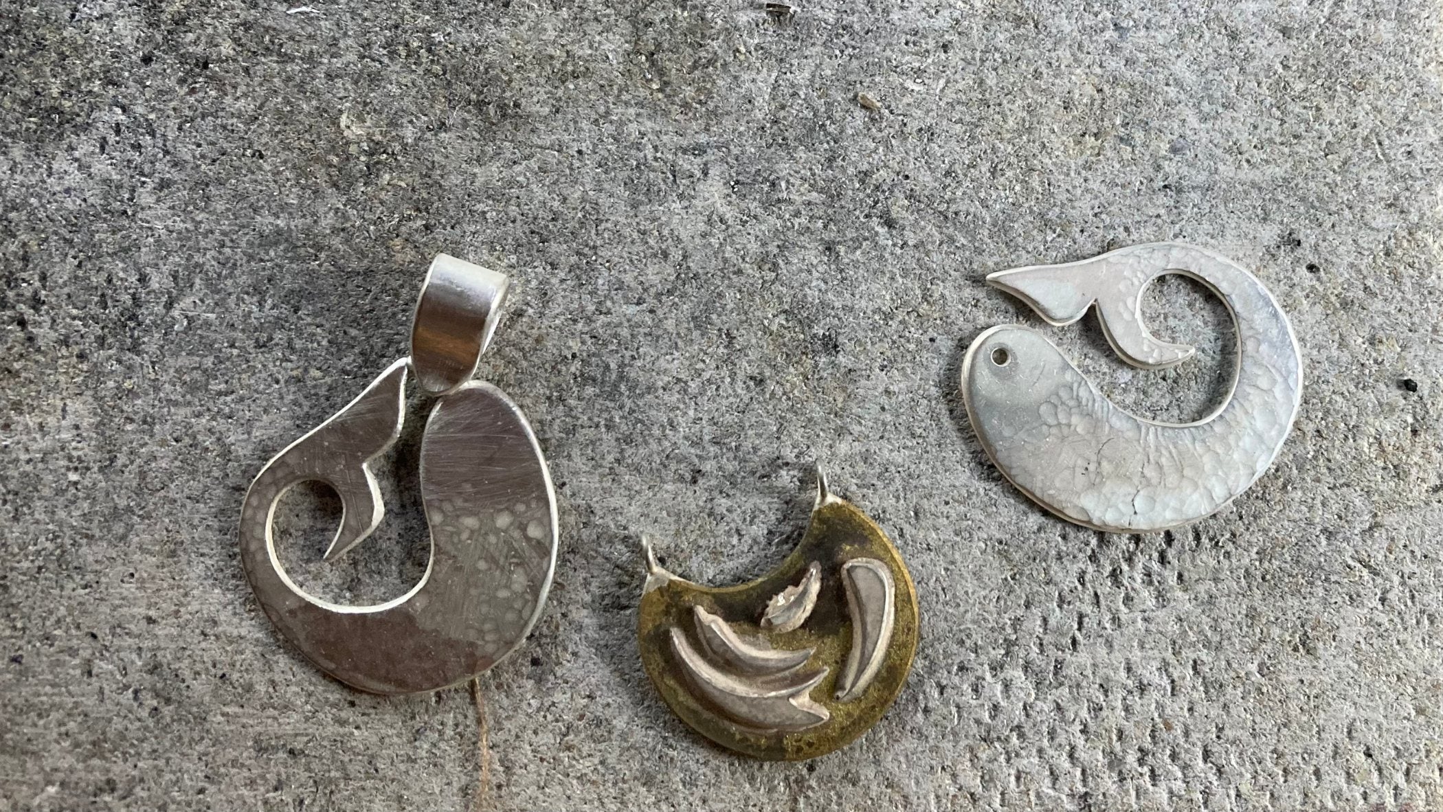 2 half-finished sterling silver fish pendants, on a heat-proof tile, with a brass and silver 'firework' necklace pendant in the middle