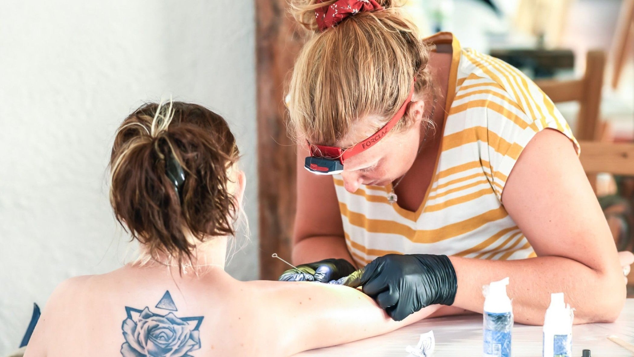 Awina in action with torch and gloves doing a handpoke tattoo on a girl's arm at a table PHOTO CREDIT: Createconnect@agency
