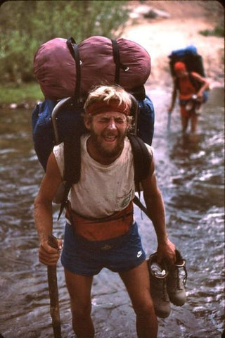 Man with backpack and bandana crossing creek, making a funny face