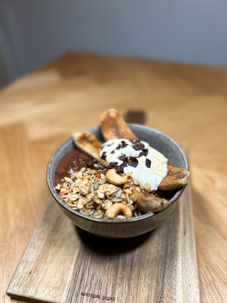 Havregrød med bagt banan og Nutella topping serveret i en keramikskål, drysset med Danish Granola Company granola, cashewnødder og mørke chokoladestykker på et træbord.