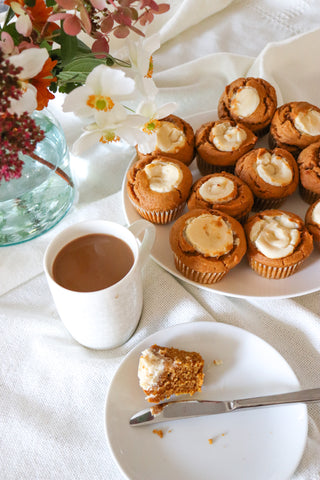 cutting into a pumpkin cream cheese muffin with a tray of pumpkin muffins, a coffee, and fresh flowers.