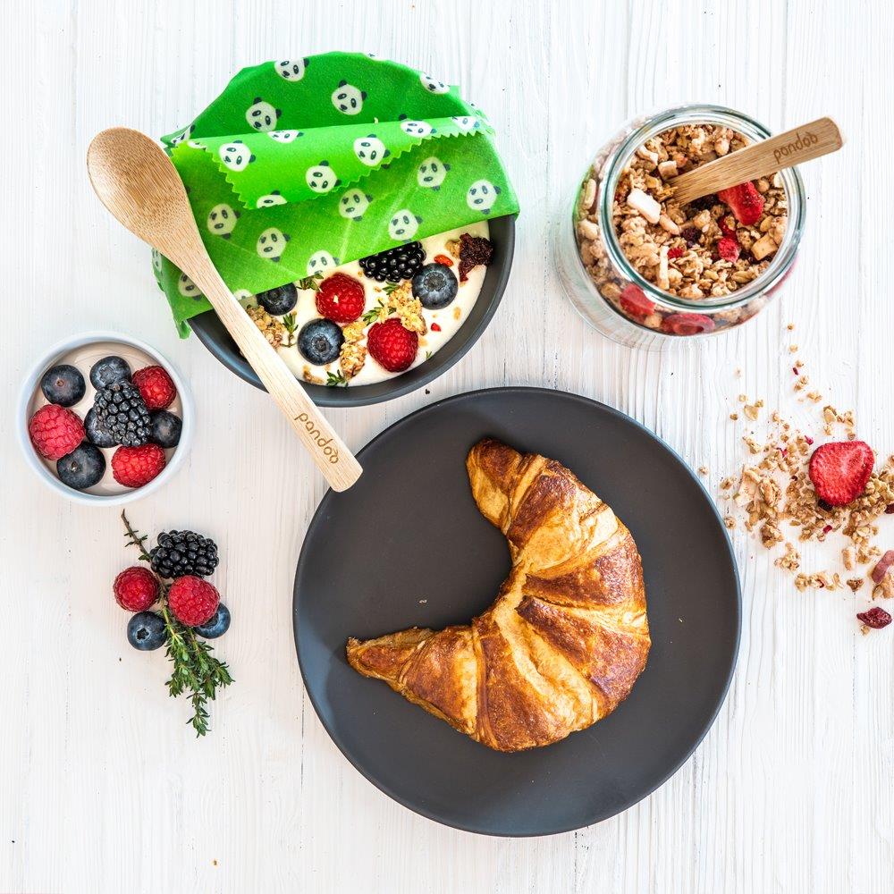 Ein gedeckter Frühstückstisch mit Croissant und einer Schale farbenfrohem Wildbeeren-Müsli. Die Schale ist halb abgedeckt mit grünem Bienenwachspapier