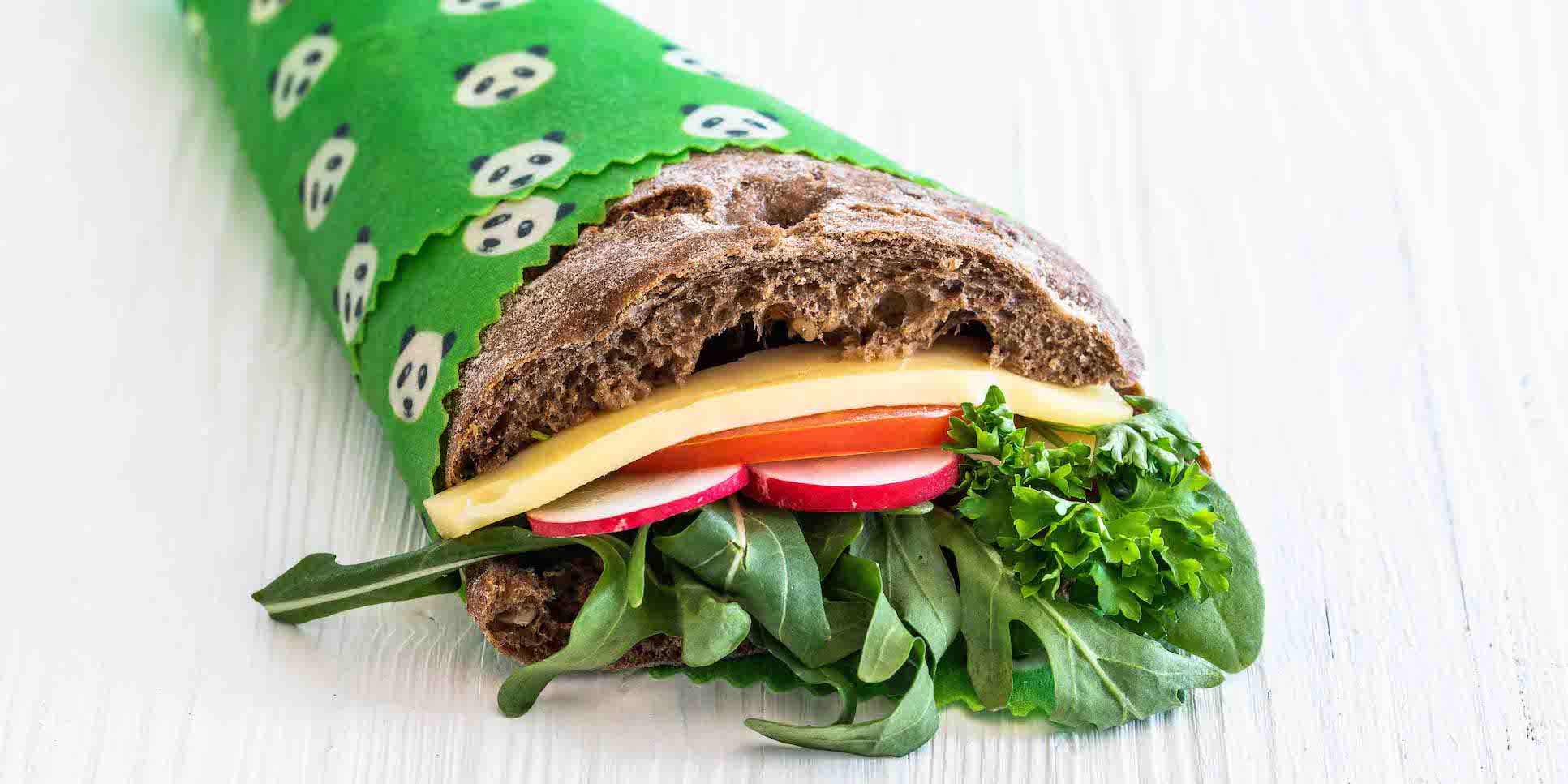 Nahaufnahme von einem dunklen Vollkorn-Sandwich belegt mit Salat, Käse, Radieschen und Rucula. Das Brot ist eingewickelt in grünem Bienenwachspapier mit Pandas darauf