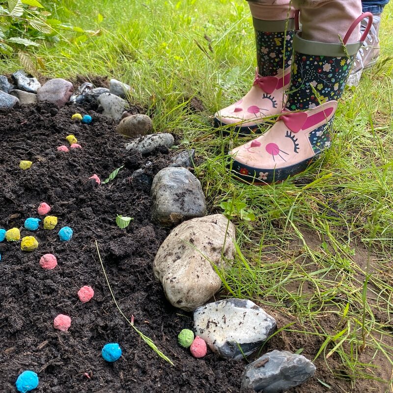 Kinderstiefel stehen neben einer frisch gesäten Reihe bunter Samenbälle im Gartenboden.