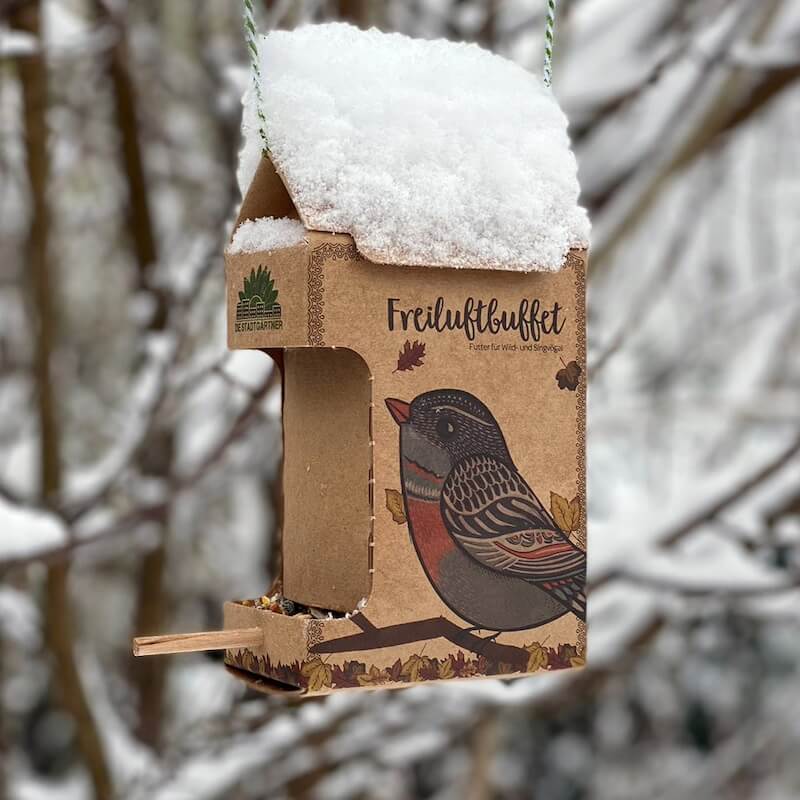 Ein Vogelfutterhaus mit Schneedecke und einem aufgedruckten Vogel an einem winterlichen Tag.