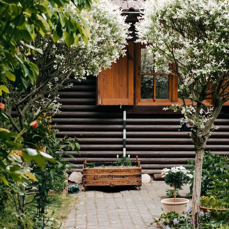 Eine idyllische Gartenszene mit einem hölzernen Blumenkasten vor einem Blockhaus und blühenden Bäumen.
