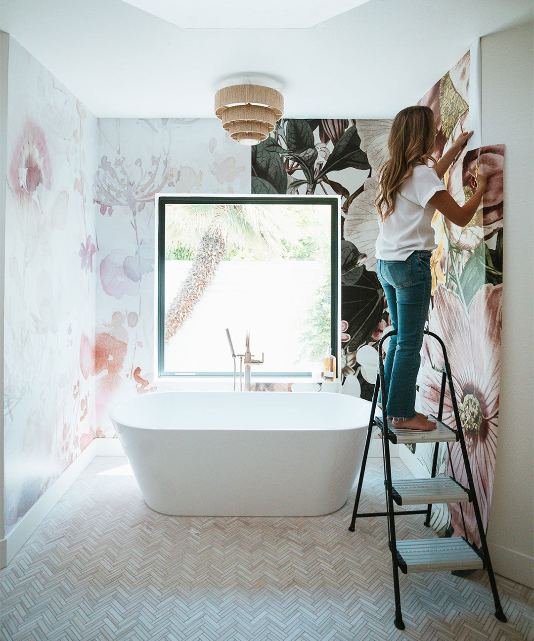 A woman is installing a dark-colored wall mural on the right side of a bright, white bathroom. On the left side of the bathroom, a light-colored wall mural is already in place. The space is well-lit with natural light coming through a window, allowing a clear comparison between the two murals.