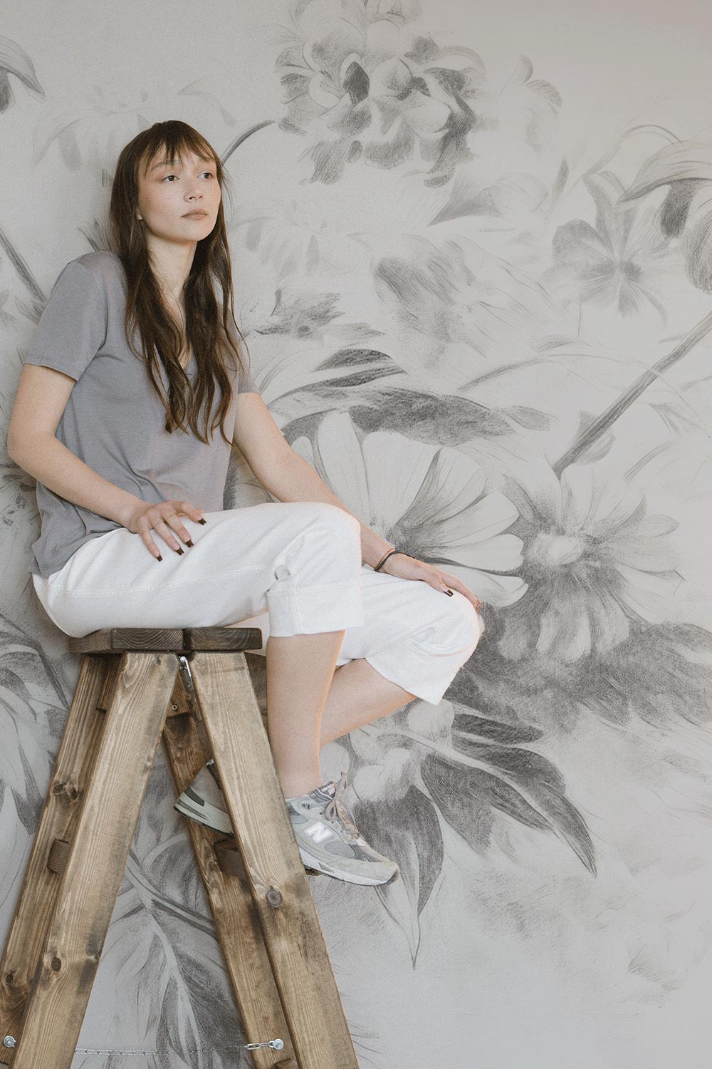 Girl sitting on a wooden ladder, in front of a greyscale sketched floral wall mural.