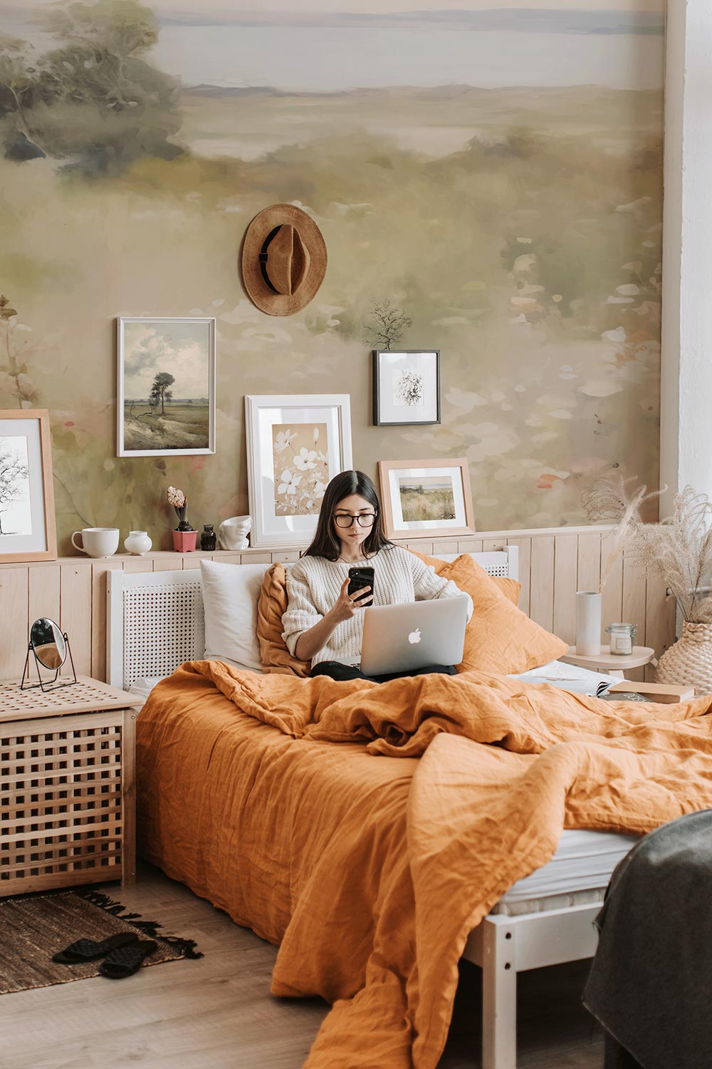 Woman sitting on bed looking at her phone. behind her there is a soft and bright floral meadow mural featured in a bedroom.