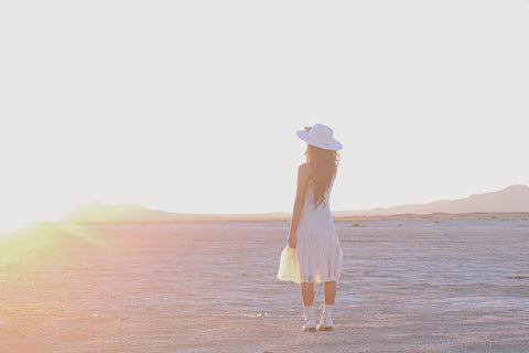 A woman in a white dress and white felt hat stands in a dry desert playa  facing away from the camera at the setting sun.
