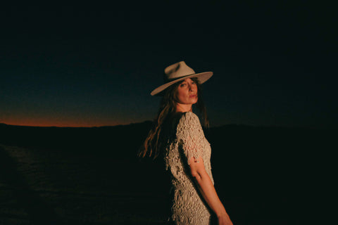 After the sun has set, a woman looks at the camera, hair swinging as she turns from the other way. She is wearing a white dress and white felt hat.