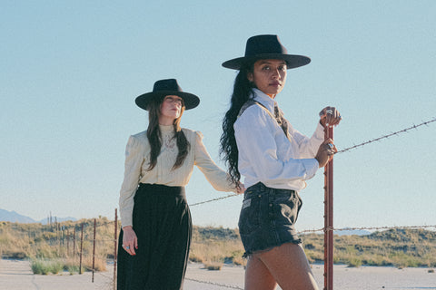 Two women with long dark hair stand at a barbed wire fence in a dry desert playa. The one in front is looking at the camera and wears a western white long sleeve shirt and black denim shorts with a navy felt hat. The woman next to her is wearing the same hat with a cream and black dress and looks off into the distance.